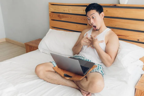 Sorprendido y sorprendido hombre está trabajando con su computadora portátil en su acogedora cama. Concepto de estilo de vida freelancer exitoso . — Foto de Stock