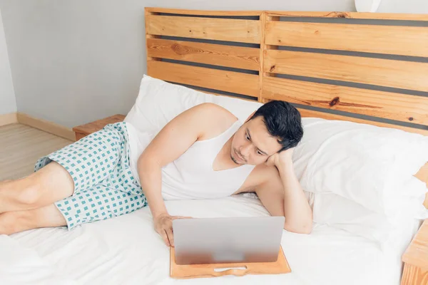 Sleepy hombre está trabajando con su portátil en su acogedora cama. Concepto de aburrido estilo de vida freelancer . — Foto de Stock