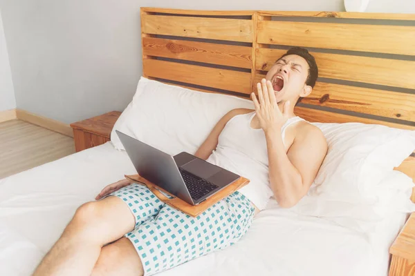 Sleepy hombre está trabajando con su portátil en su acogedora cama. Concepto de aburrido estilo de vida freelancer . — Foto de Stock