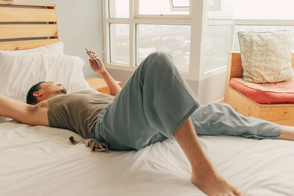 Asiático hombre mentira en cama y relajarse en su apartamento en calidez domingo verano primavera . — Foto de Stock