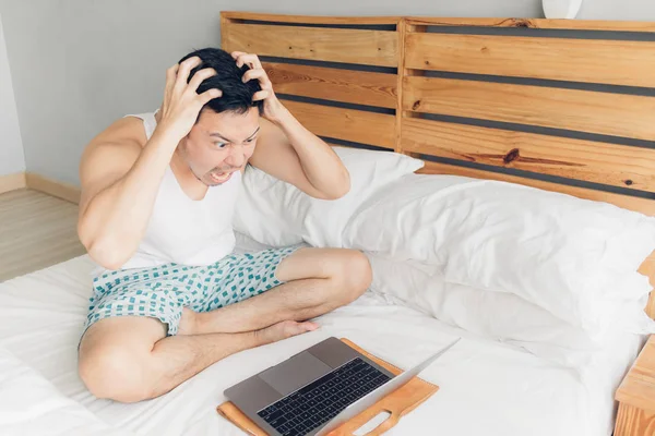 Angry man is working with his laptop on his bed. Concept of freelancer problems.