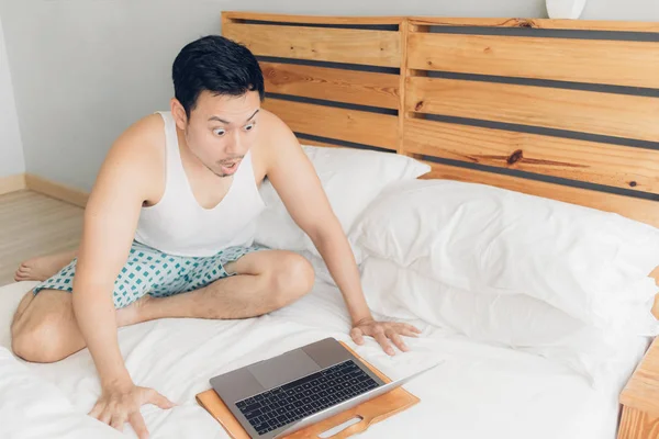 Homem surpreso e chocado está trabalhando com seu laptop em sua cama aconchegante. Conceito de estilo de vida freelancer de sucesso . — Fotografia de Stock