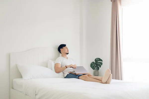 Pria yang berbaring di tempat tidur mengerjakan laptopnya di kamar tidur putih yang nyaman. — Stok Foto