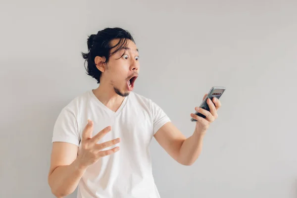 Surpreso e chocado homem de cabelo longo em t-shirt branca está usando smartphone. — Fotografia de Stock