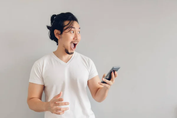 Surpreso e chocado homem de cabelo longo em t-shirt branca está usando smartphone. — Fotografia de Stock