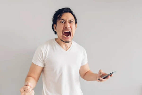 Irritado e furioso homem de cabelo longo em t-shirt branca está usando smartphone. — Fotografia de Stock