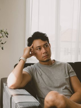 Kısa saçlı Taylandlı bir adam, temiz, modern bir odada gri bir kanepeye oturmuş, gün ışığında, minimalist bir evde, perdenin yanında sessizce düşünürken, yan görüntüden yakalanmış..