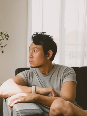 Gri tişörtlü Taylandlı adam kollarını kavuşturmuş, parlak, minimalist oturma odası ortamında düşünceli bir ifadeyle sola bakıyor.