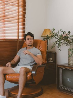 Kavisli bir şezlonga yaslanmış kısa saçlı Taylandlı adam. Güneş ışığı ve jaluziler, lamba ve ahşap büfe de dahil olmak üzere retro ev dekorasyonu..