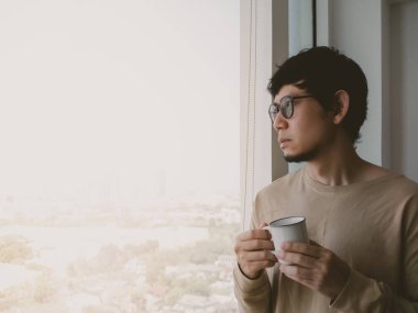 Adam bir fincan kahve tutuyor ve pencereden dışarı bakıyor. Sahne, ufuk çizgisi manzaralı bir şehirde geçiyor. Adam düşüncelere dalmış, muhtemelen gününü ya da çevresini düşünüyor.