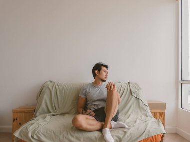 Taylandlı erkek, gri gömlekli yatakta çaprazlama oturuyor ve sakin, minimalist yatak odasına bakıyor, Asya kökenli.