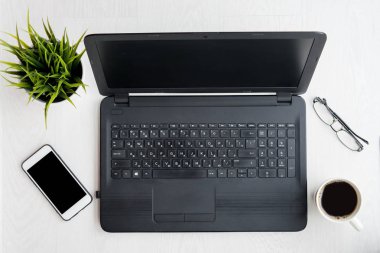 Dizüstü bilgisayarı, dizüstü bilgisayarı, telefonu, bardakları, kahvesi ve bilgisayarı siyah ekranlı bitkisi olan modern beyaz masa. Düz yatıyordu