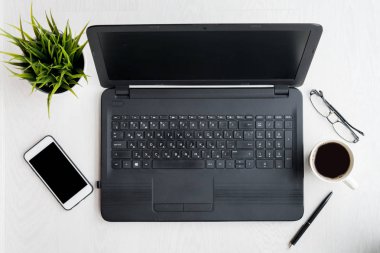 Laptop bilgisayarı, dizüstü bilgisayarı, telefonu, gözlükleri, kalemi, kahvesi ve siyah ekranlı bitkisi olan modern beyaz masa. Düz yatıyordu