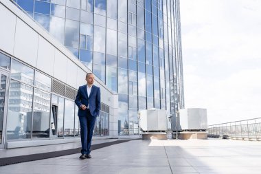 Takım elbiseli ve beyaz gömlekli genç bir adam büyük bir ofis merkezinin yanında yürüyor. Modern bir iş merkezinin yakınındaki bir şehirde iş adamı. Büyük, cam bir binanın yakınındaki bir işadamı.