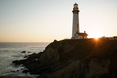 Okyanusta gün batımına karşı arka planda beyaz bir deniz feneri ve küçük eski bir ev. Yeşil çimenleri ve güçlü dalgaları olan kayalık sahil, gün batımından önce taşlarda beyaz bir köpük oluşturur. Gün sonunda yaz güneşiyle birlikte.