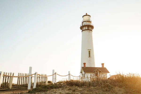 カリフォルニア州の太平洋岸にある古い白い灯台とその近くの建物 遠くの船や船に輝く夜を待つ灯台 — ストック写真