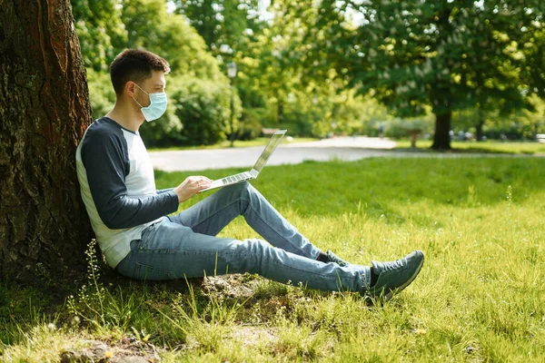 A handsome young man in a medical mask sits in a park and works on a laptop under a green tree on a summer street background. Masked guy working on a computer in the park