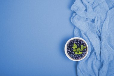 Fresh wild blueberries with green leaves in a bowl on blue background. Top view. clipart