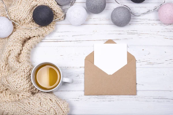 Té de la taza, cuadros beige, tarjeta en blanco en sobre de artesanía abierta, decorado con bolas de algodón guirnalda ligera sobre fondo de madera blanca . —  Fotos de Stock