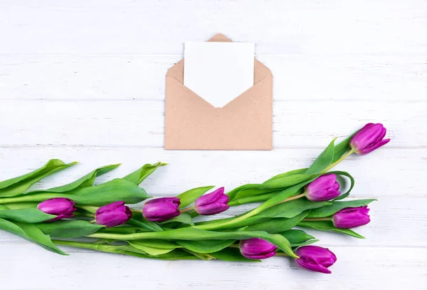 Композиція білої порожньої картки у відкритому ремісничому конверті, прикрашена свіжими красивими фіолетовими тюльпанами на білому дерев'яному фоні з місцем для тексту . — стокове фото