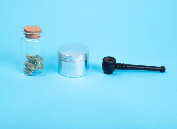 Brotes de cannabis medicinal seco en una botella de vidrio, molinillo de plata y tubo de madera sobre fondo azul . — Foto de Stock