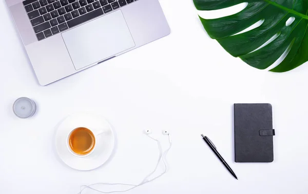 Office workspace with laptop, tropical leaf Monstera, notebook, candle, earphones and cup of coffee on white background.