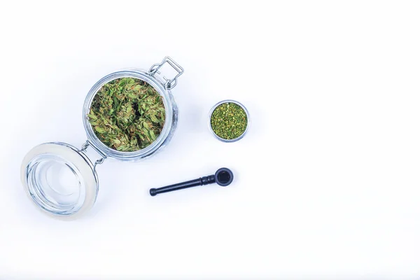 Fresh green buds or flowers of cannabis marijuana in opened transparent glass jar and grinded weed in open aluminum round metal box and smoking pipe isolated on white background. — Stock Photo, Image