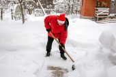 muž v červených dolů sako a red hat Santa Claus vymaže sníh na dvorku. Vymaže závěje na cestu k domovu. Po husté sněžení. Člověk odstranit sníh s velkými plastová lopata s dřevěnou rukojetí