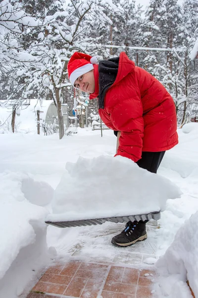 man in red down jacket and red hat of Santa claus clears snow in backyard. Clears snowdrifts on path to home. In snowfall. Man remove snow with big plastic shovel with wooden handle