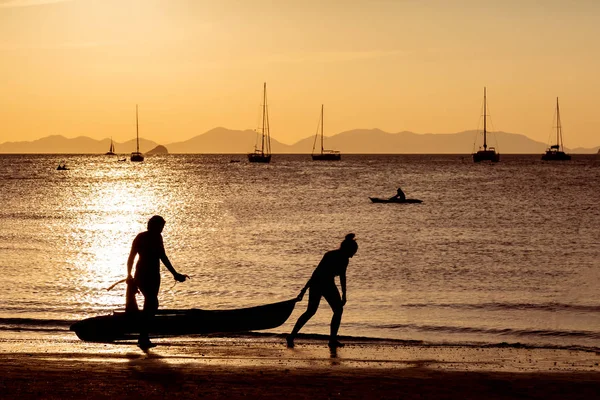 People on the beach play sports in the light of a golden sunset. Kayakers float in the water, two girls carry out to the land of the boat. Against the background of the yacht and the mountains, the reflection of the sun on the water, the path.