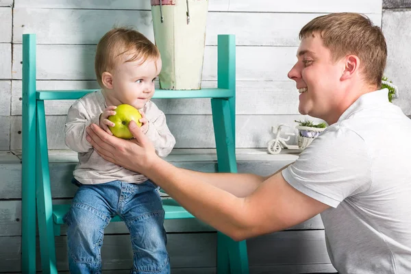 Father gives to baby boy big green apple. He both are in jeans and white hoodie.  Dad with son sits on the steps indoor. White wooden background