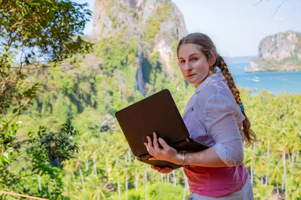 Woman in white shirt with black laptop on the top of hill. Remote freelance working, touristic routes creating. View on the sea, mountains and tropical jungles at sunny day