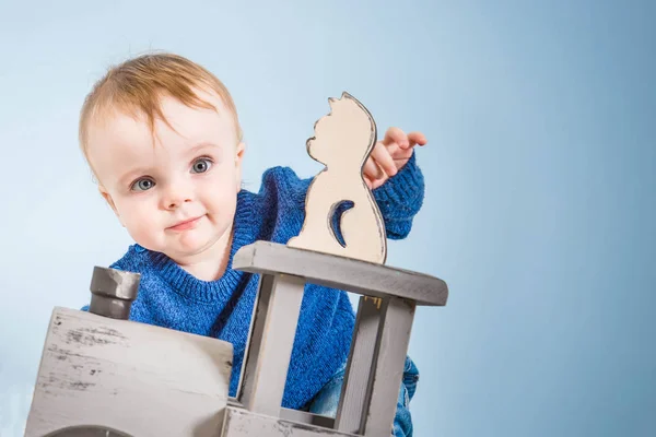 Happy one year old child in a blue sweater plays wooden toys. Blonde baby boy on blue background. Cat and train of wood