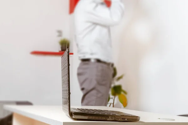 Laptop on the table in the office. Against a background in blur, a young businessman is talking on the phone and looking at the wall