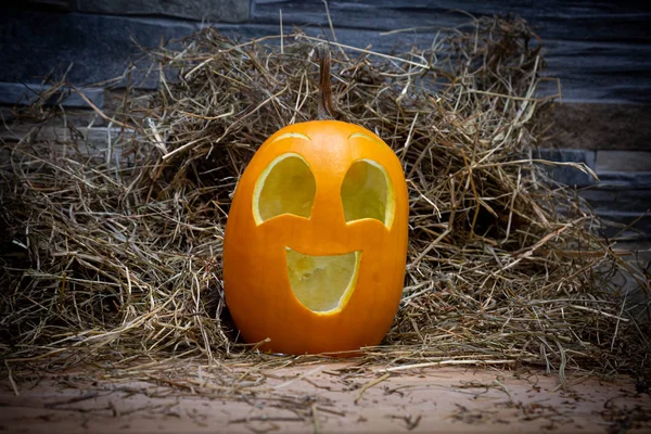 Yellow and happy smiling pumpkin. Halloween symbol on a gray stone wall background, stands on a hay and a wooden stand. Jack o lantern