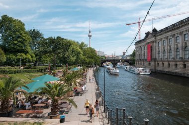 Berlin, Almanya - Haziran 2018: Arka planda turist tekneleri ve tv kulesi ile Spree Nehri'nin yanındaki plaj barında insanlar