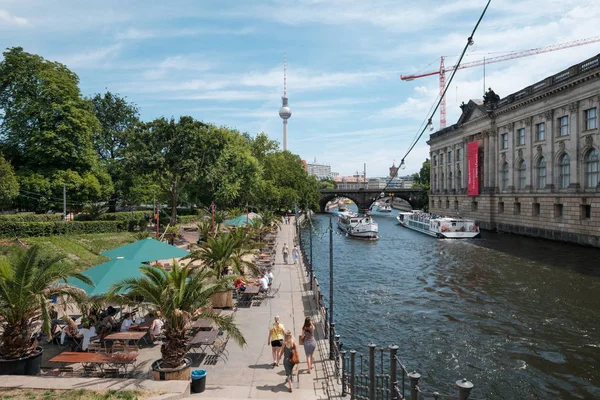 Berlin, Almanya - Haziran 2018: Arka planda turist tekneleri ve tv kulesi ile Spree Nehri'nin yanındaki plaj barında insanlar