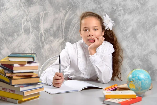 The schoolgirl in school uniform sits at the desk and thinks about the decision of the task on a light gray background. Back to school. The new school year. Child education concept.