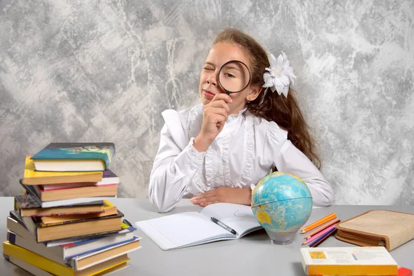 The schoolgirl in school uniform sitting and looking at us through a magnifying glass on a light gray background. Back to school. The new school year. Child education concept.