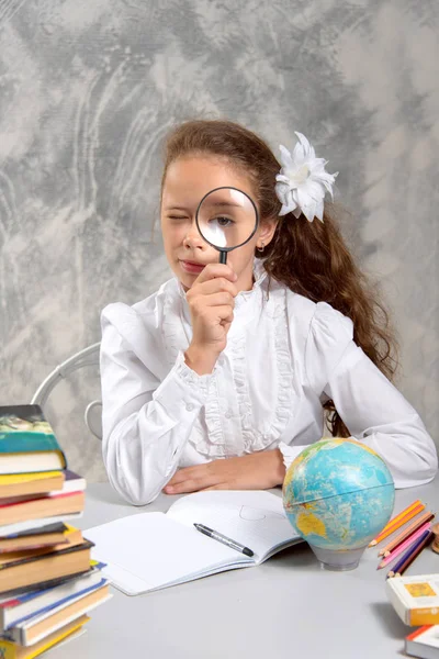 The schoolgirl in school uniform sitting and looking at us through a magnifying glass on a light gray background. Back to school. The new school year. Child education concept.