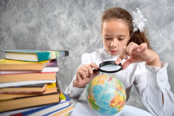 The schoolgirl in school uniform looking at a globe through a magnifying glass and writing something in a notebook. Back to school. The new school year. Child education concept. A geography lesson