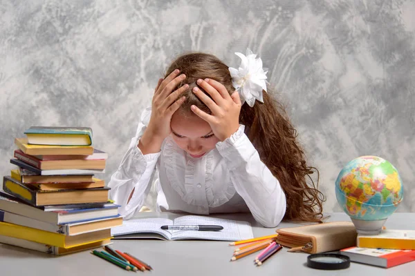 The schoolgirl in school uniform sits at the desk and thinks hard about the decision of the difficult task on a light gray background. Back to school. The new school year. Child education concept.