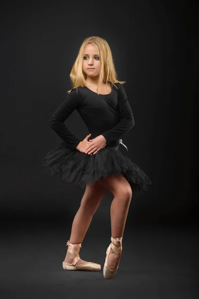 Милая маленькая балерина с длинными волосами в черной пачке и пуантом, позирующая на черном фоне — стоковое фото