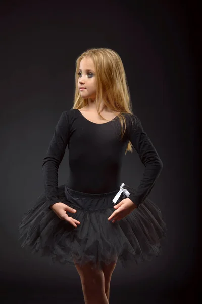 Милая маленькая балерина с длинными волосами в черной пачке и пуантом, позирующая на черном фоне — стоковое фото