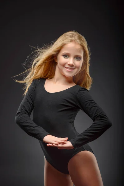 Милая маленькая балерина с длинными волосами в черном трико и пуантом, позирующая на черном фоне — стоковое фото