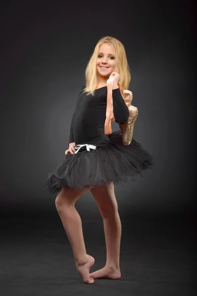 Cute little ballerina with long hair in a black tutu and pointe posing on a black background