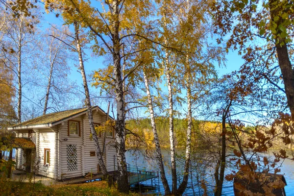 这座古老的木制白色房子坐落在美丽的蓝色湖畔的一个黄色的秋天森林里 — 图库照片