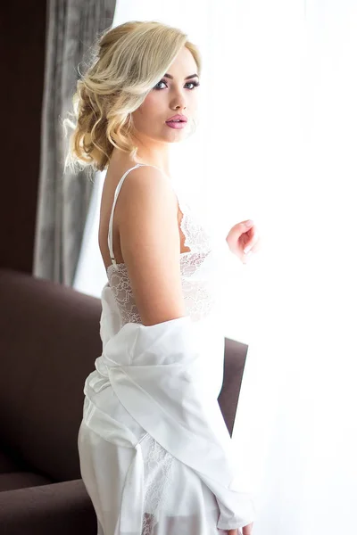 Krásná blond dívka sedí na pohovce oblečený v bílé negližé drží svatební kytici, posílání vzduchu polibek v pozadí mnoha barev — Stock fotografie