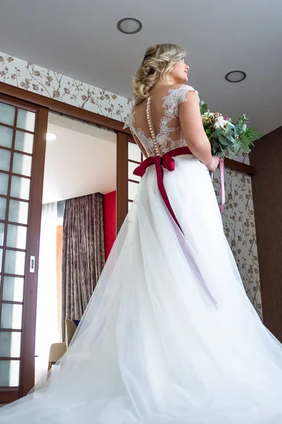 beautiful young girl with make-up and hairdo stands in a hotel room in a wedding dress with a red ribbon and with a wedding bouquet in her hands.