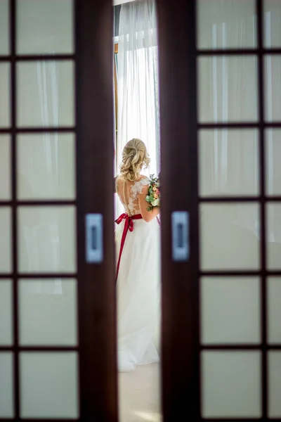 a girl in a wedding dress with make-up and hairdo stands in a bright hotel room near the window and looks out into the street in a wedding dress with a red ribbon and with a wedding bouquet in her hands.